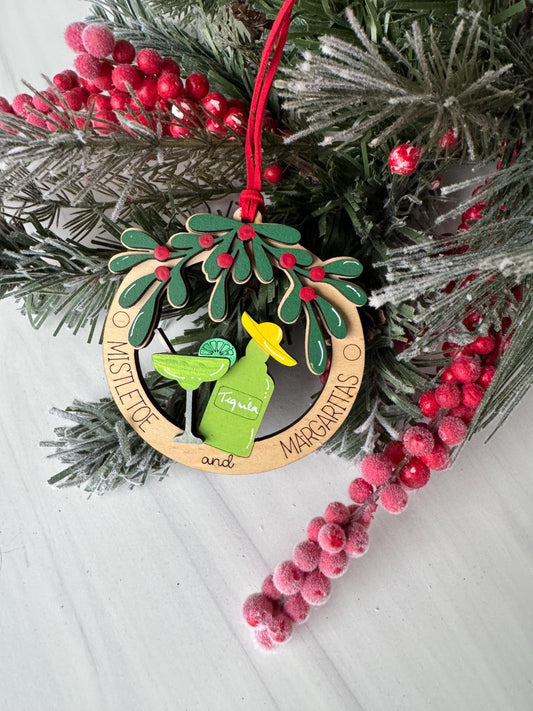A Christmas ornament with a green bottle and a yellow hat, surrounded by a wreath of greenery and red berries, placed on a white surface.