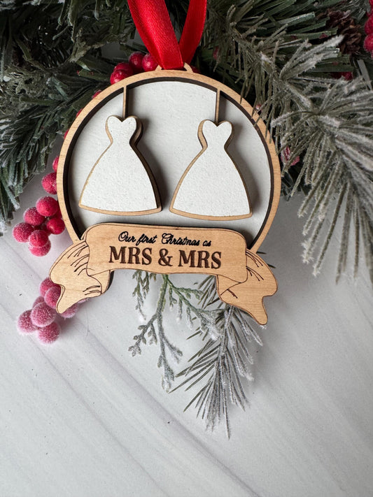 A wooden ornament with a ribbon and two white dress shapes, placed on a white surface with pine branches and red berries.