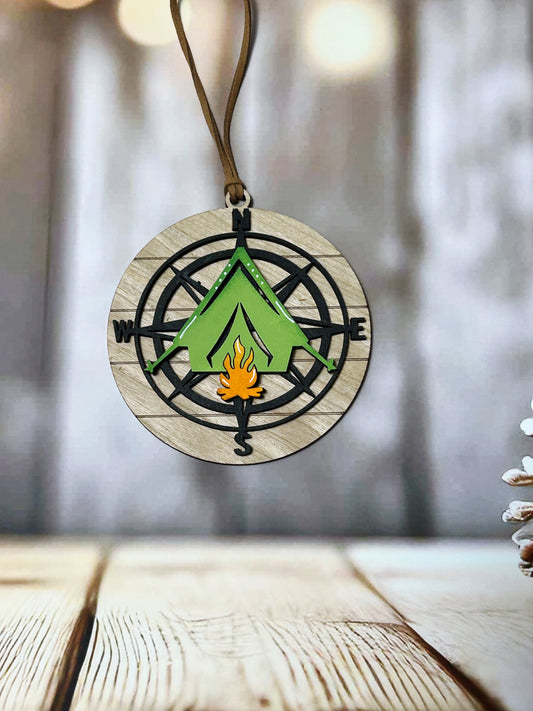 A wooden compass pendant with a green tent design and a fire symbol, hanging on a string against a blurred background.