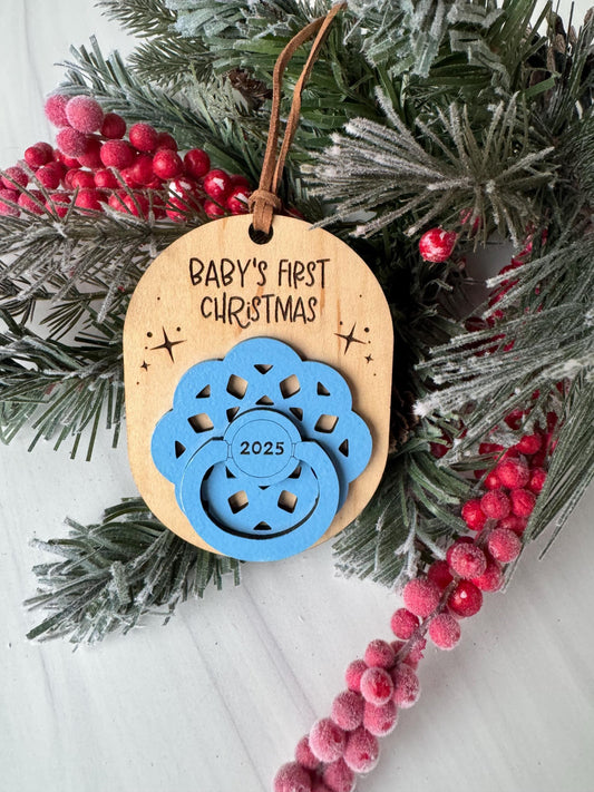 A wooden ornament with a blue design and the text "BABY'S FIRST CHRISTMAS" is displayed on a Christmas tree branch, surrounded by pine branches and red berries.