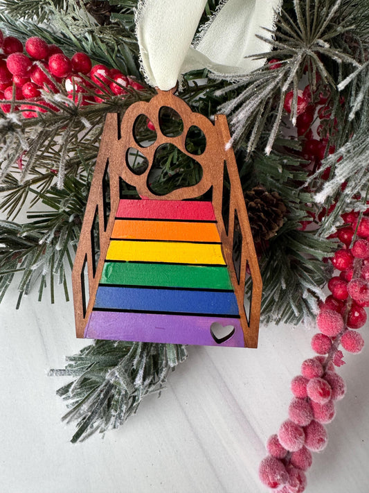 A wooden ornament with a rainbow-colored design is displayed on a Christmas tree, surrounded by pine branches, berries, and a white ribbon.