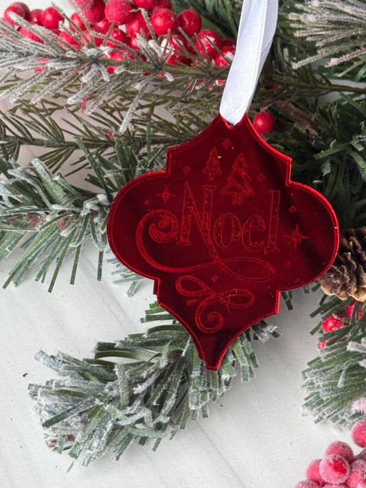 A red ornament with the word "Noel" engraved on it, hanging on a Christmas tree branch surrounded by pine needles and red berries.