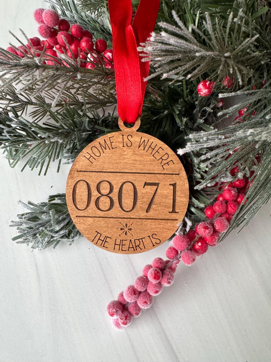 A wooden ornament with the text "HOME IS WHERE THE HEART IS" carved into it, hanging on a pine branch with red berries.