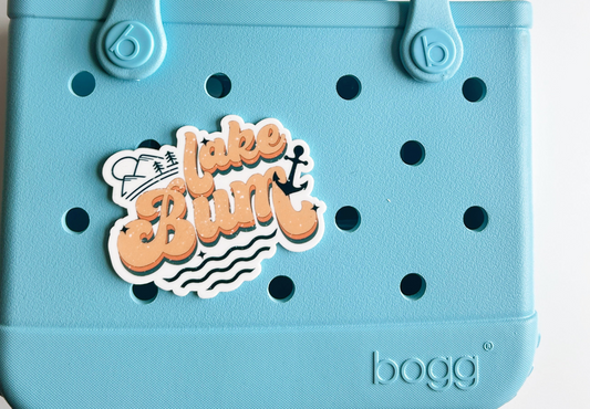 A light blue tote bag with a printed sticker that says "Lake Bum" and has a wave design.