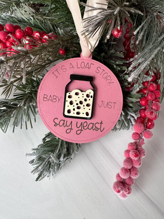 A pink ornament with a white design and text, surrounded by pine branches and red berries, placed on a white surface.