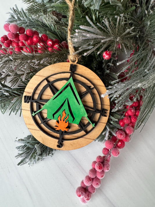A wooden ornament with a compass design and a flame-like element is displayed on a branch of a Christmas tree adorned with red berries.
