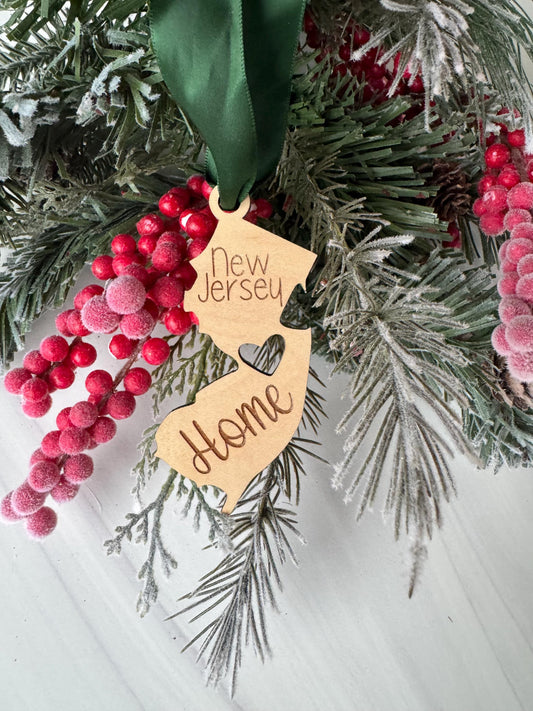 A wooden ornament shaped like the state of New Jersey, with the word "New Jersey" written on it, is hanging from a pine branch with red berries.