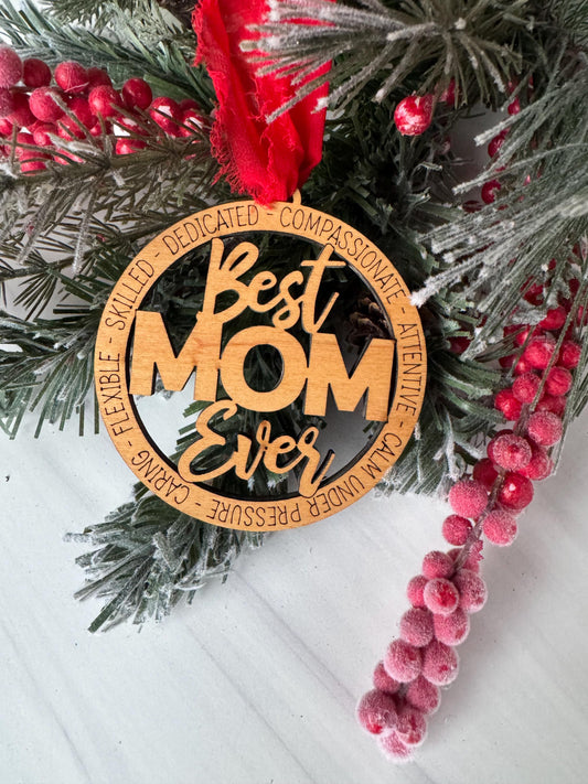 A wooden ornament with the text "Best Mom" carved into it, surrounded by pine branches and red berries, placed on a white surface.