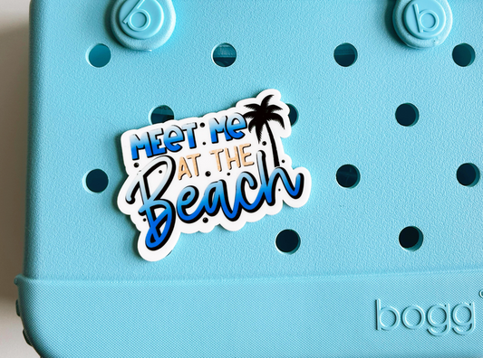 A light blue tote bag with a sticker that says "Meet me at the beach" and has a palm tree design.