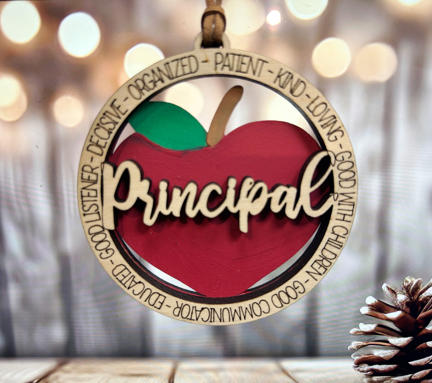 A wooden ornament in the shape of an apple, with the word "Principal" written on it, hangs against a blurred background with bokeh lighting.