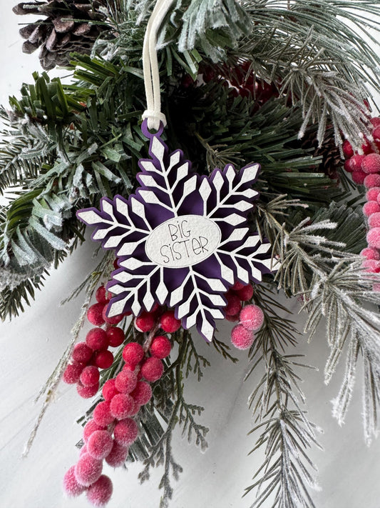 A purple snowflake ornament with the word "BIG SIS" written on it, surrounded by pine branches, berries, and pine cones, creating a festive winter scene.