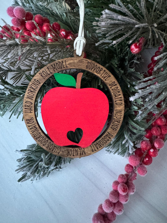 A red apple ornament with a heart-shaped cutout is displayed on a pine branch, surrounded by red berries and greenery.