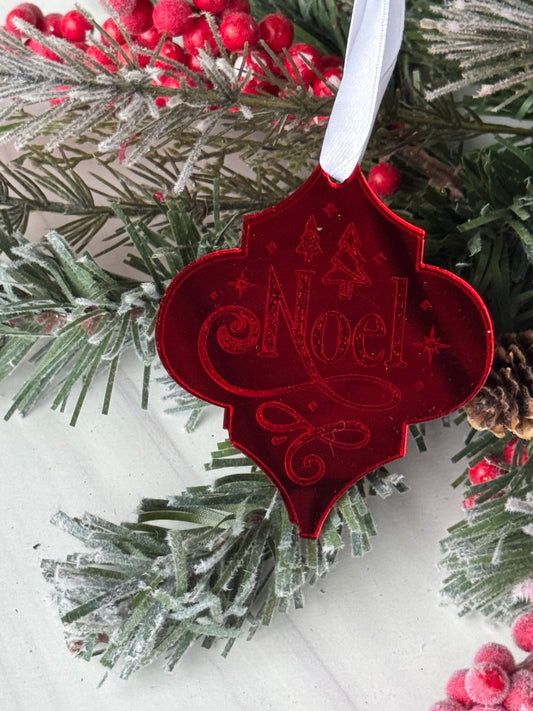A red ornament with the word "Noel" engraved on it, hanging on a Christmas tree branch surrounded by pine needles and red berries.