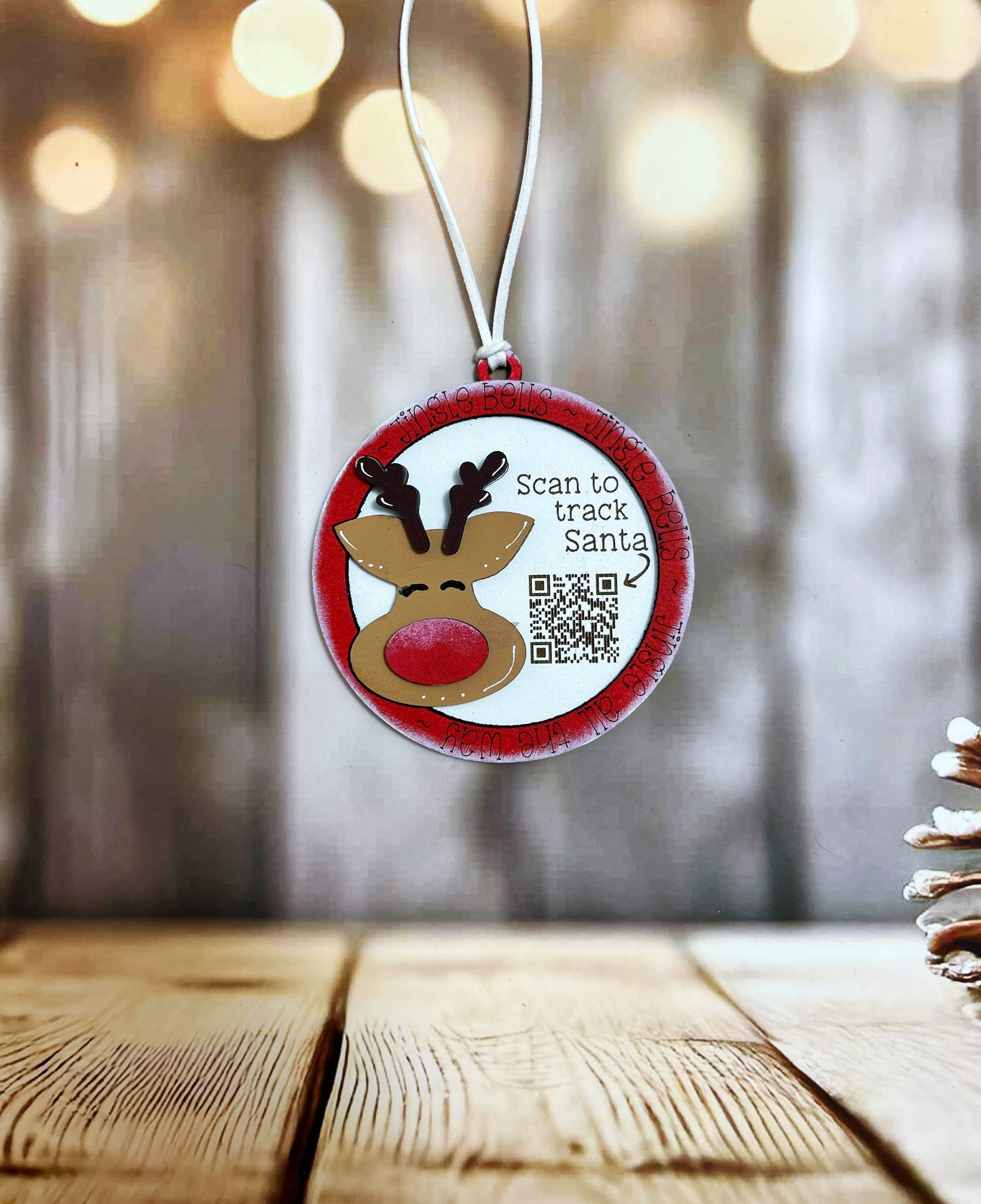 A Christmas ornament with a reindeer and the text "Scan to track Santa" is hanging on a string against a blurred background with bokeh lights.