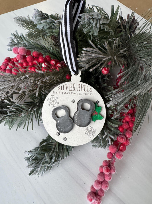 A Christmas ornament with a black and white ribbon, silver bells, and red berries is displayed on a white surface.