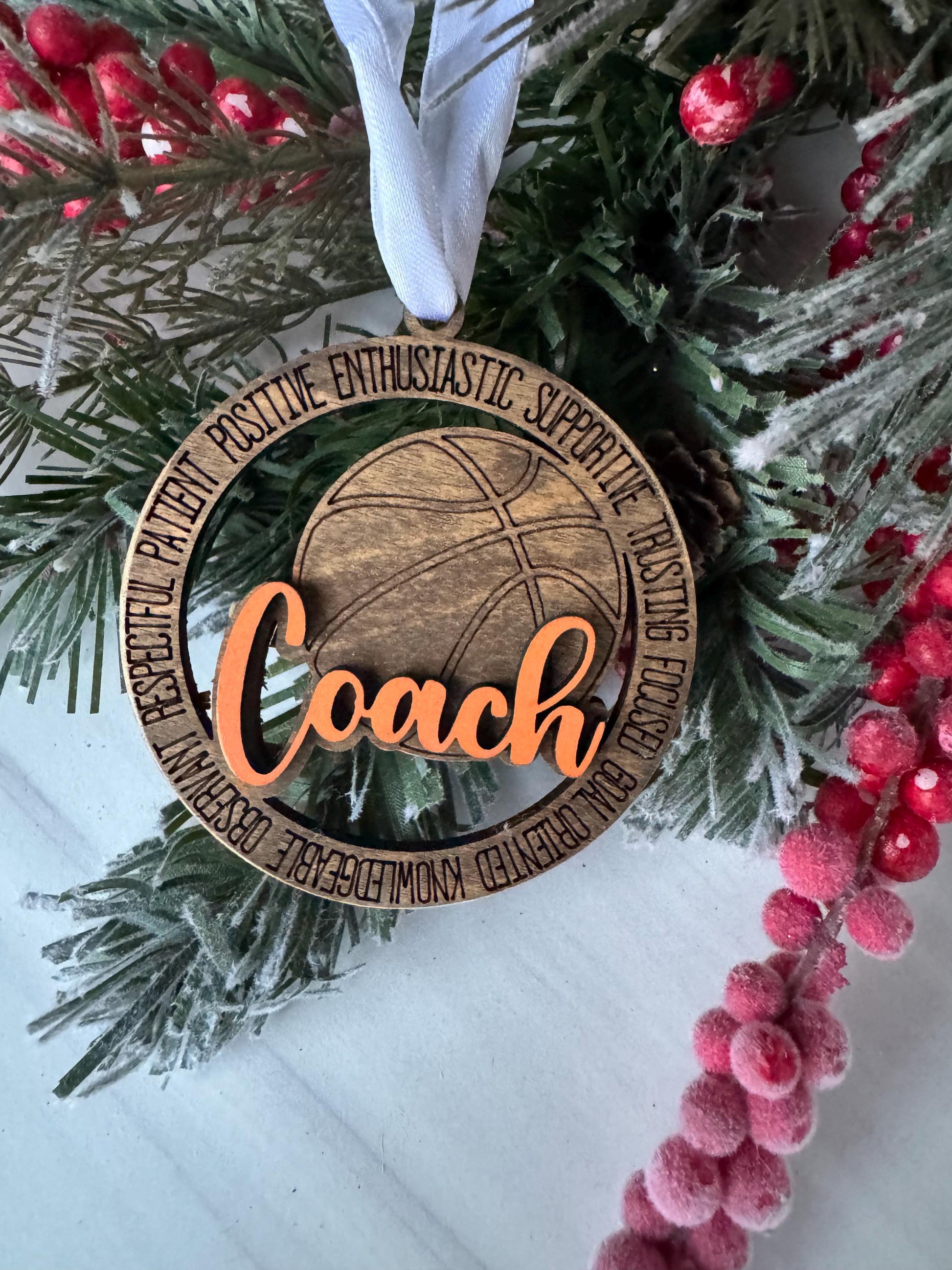 A wooden basketball ornament with the word &quot;Coach&quot; engraved on it, surrounded by a wreath of pine branches and red berries.