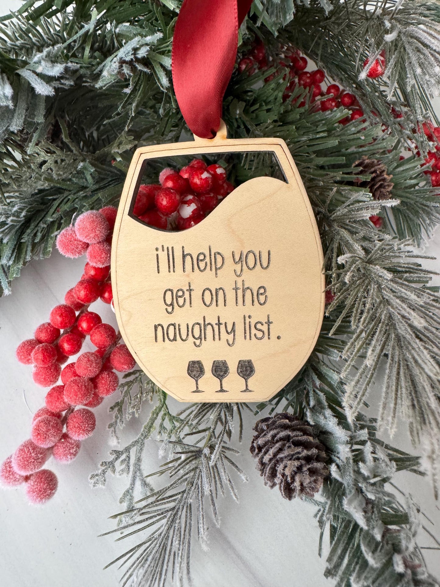 A wooden ornament with a humorous message is hanging on a wreath of pine branches and berries, surrounded by other holiday decorations.