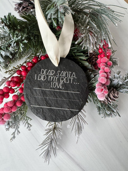 A wreath made of pine branches, berries, and pine cones, with a slate-like object featuring a message written on it, placed on a white surface.