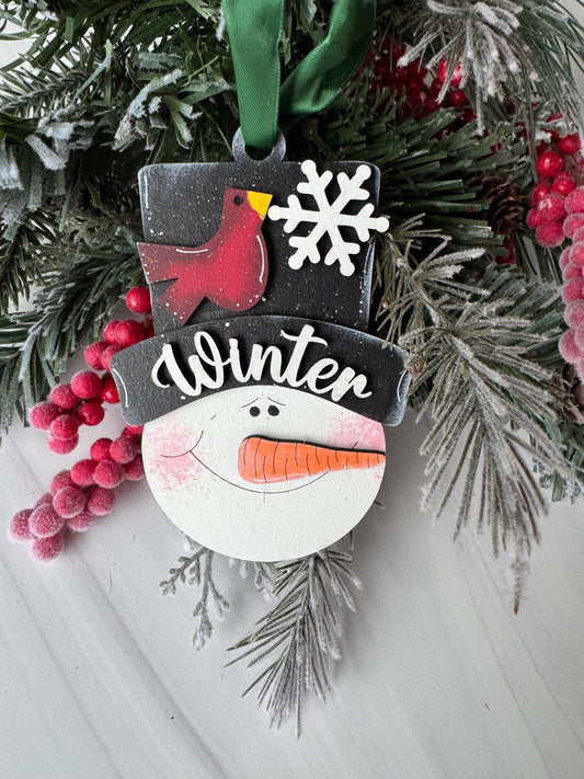 A snowman ornament with a red bird perched on top, surrounded by pine branches and red berries, is displayed on a white surface.