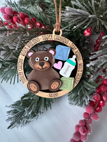 A wooden ornament with a teddy bear and baby supplies, including a bottle, is displayed on a pine branch with red berries.