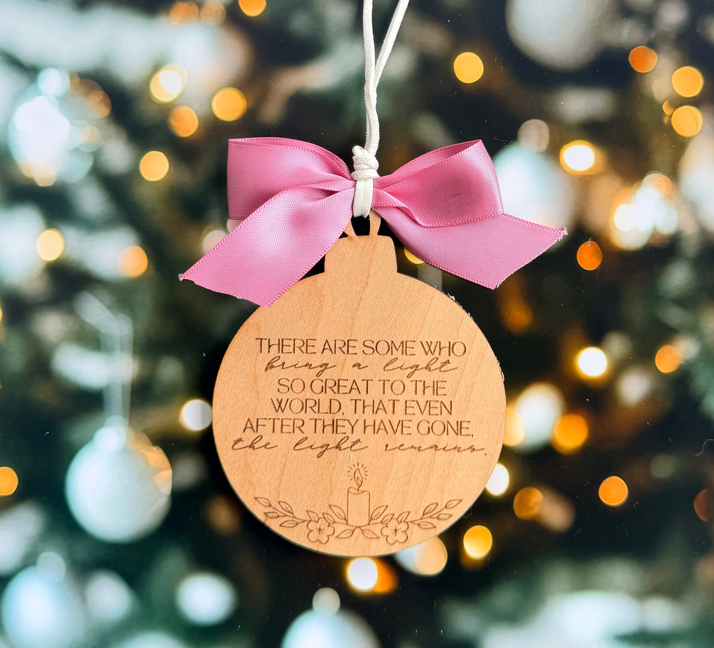 A wooden ornament with a pink ribbon and a message engraved on it, hanging against a blurred background with Christmas lights.