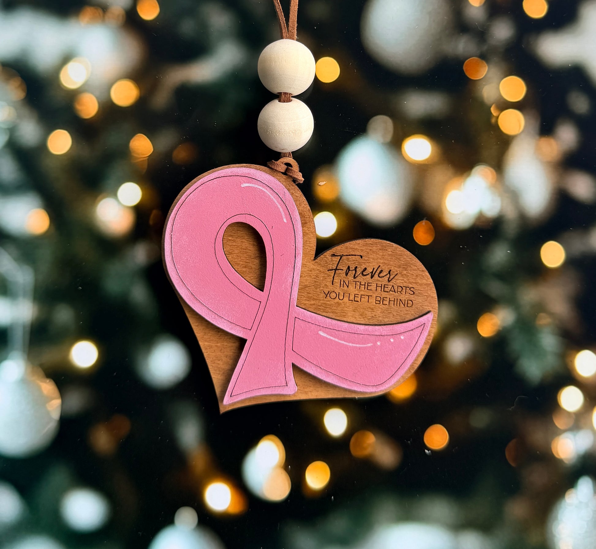 A pink ribbon ornament with the text "Forever in the hearts you left behind" is hanging against a background of blurred lights, likely representing a Christmas tree.