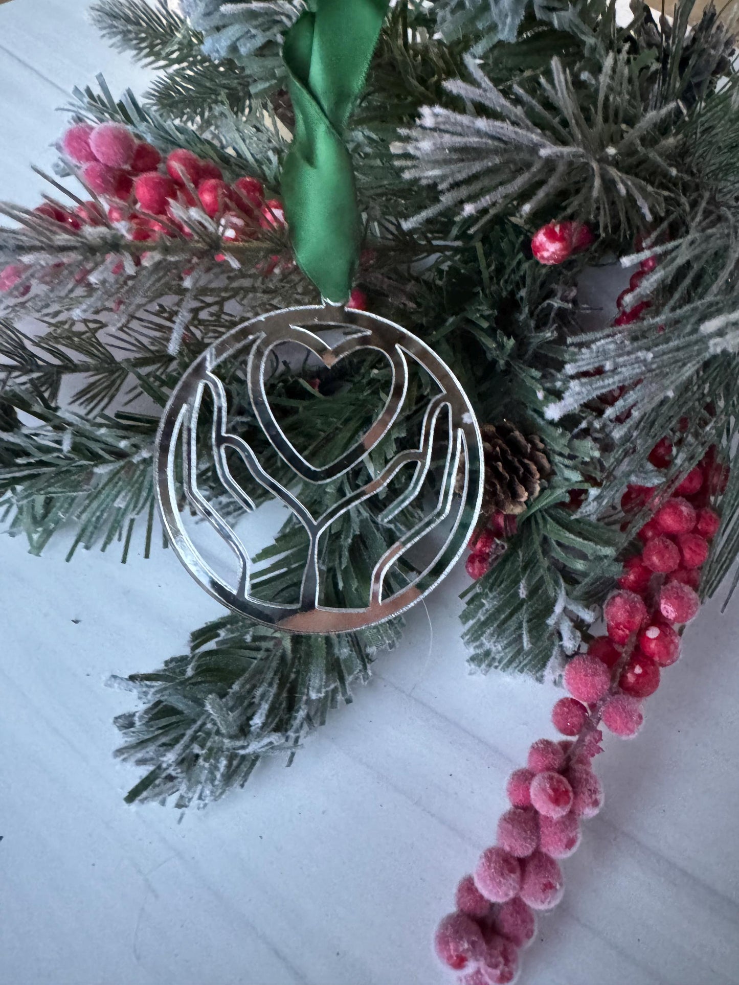 A silver ornament with a heart shape is hanging from a Christmas tree, surrounded by pine branches, red berries, and a green ribbon.