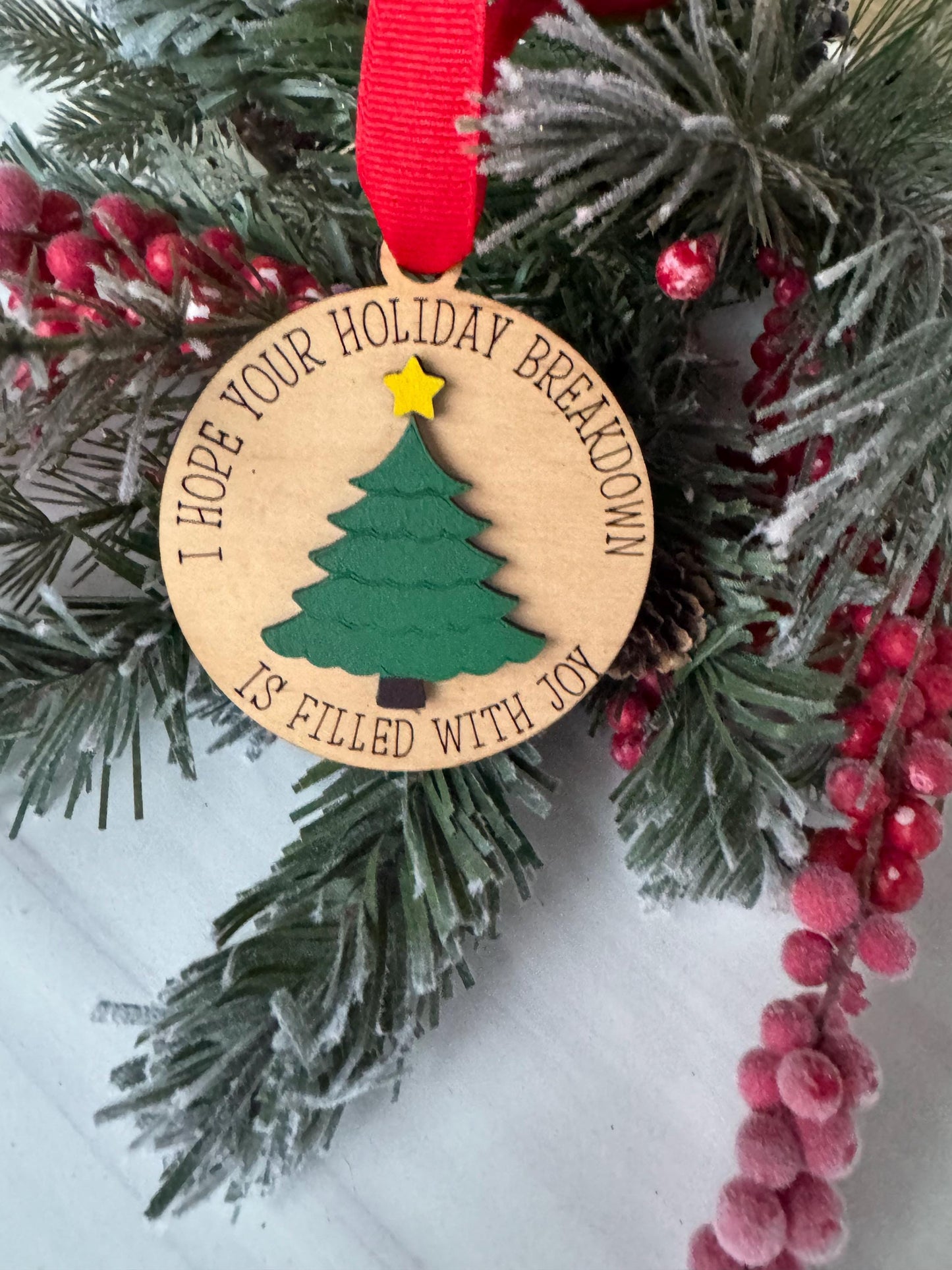A wooden ornament with a Christmas tree design, surrounded by pine branches and red berries, is displayed on a white surface.