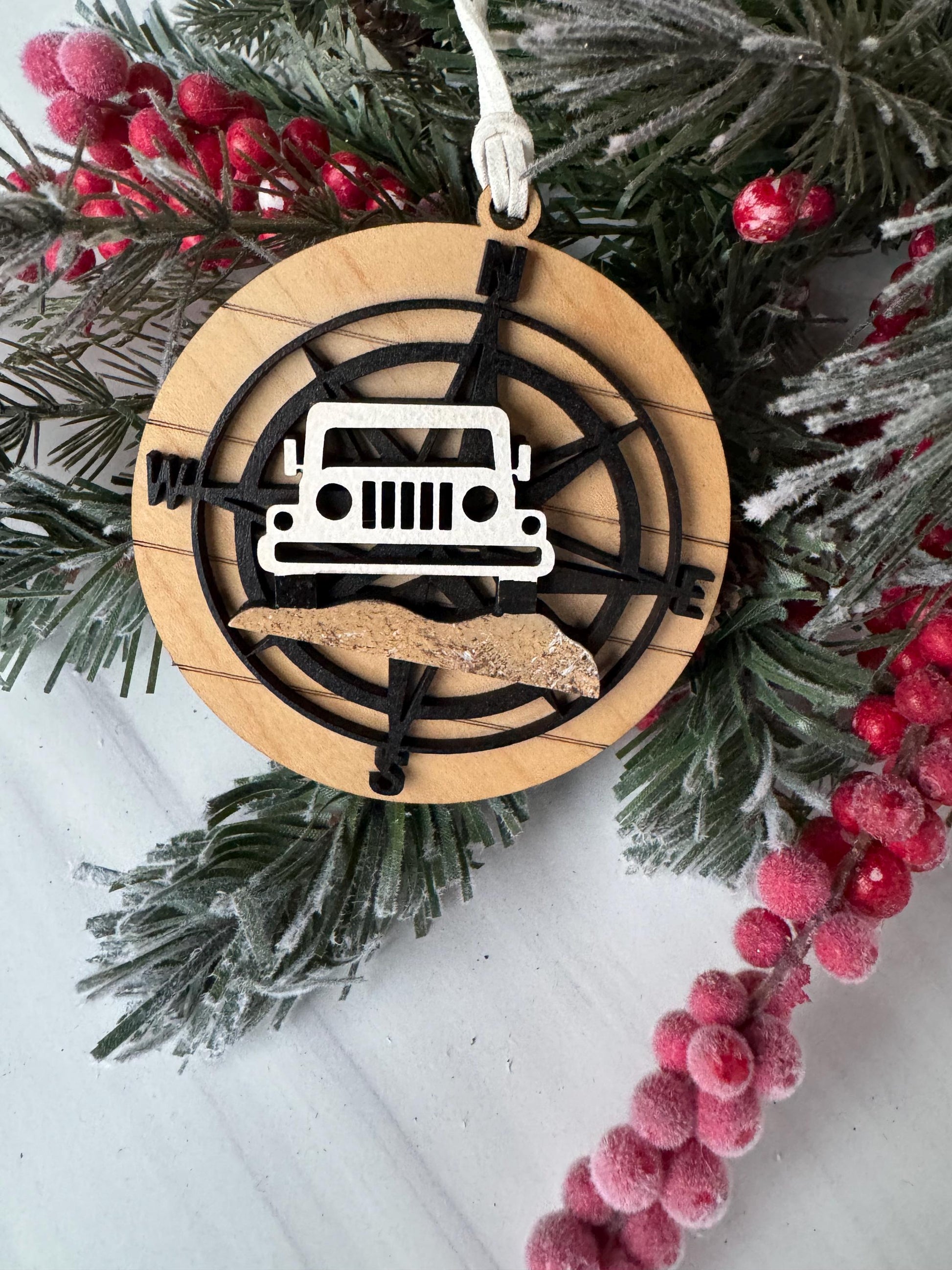 A Christmas ornament with a compass and a jeep, surrounded by pine branches and red berries.