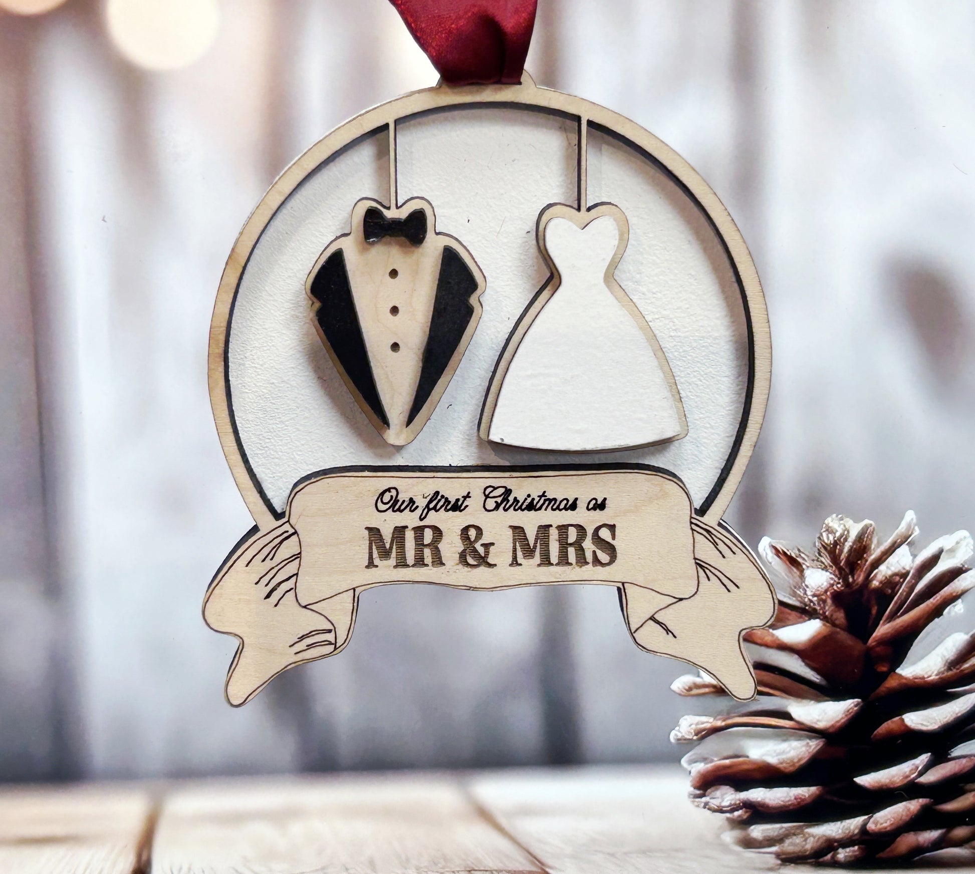 A wooden ornament with a tuxedo and a wedding dress, along with a banner that says "Our first Christmas as Mr & Mrs". The ornament is hanging from a ribbon, and there is a pine cone next to it.