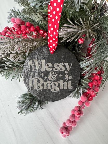 A black stone ornament with the text &quot;MESSY & BRIGHT&quot; is surrounded by a wreath of pine branches, red berries, and a red polka dot ribbon.
