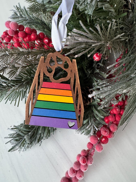 A colorful wooden ornament with a rainbow pattern is hanging on a pine branch, surrounded by red berries and greenery.