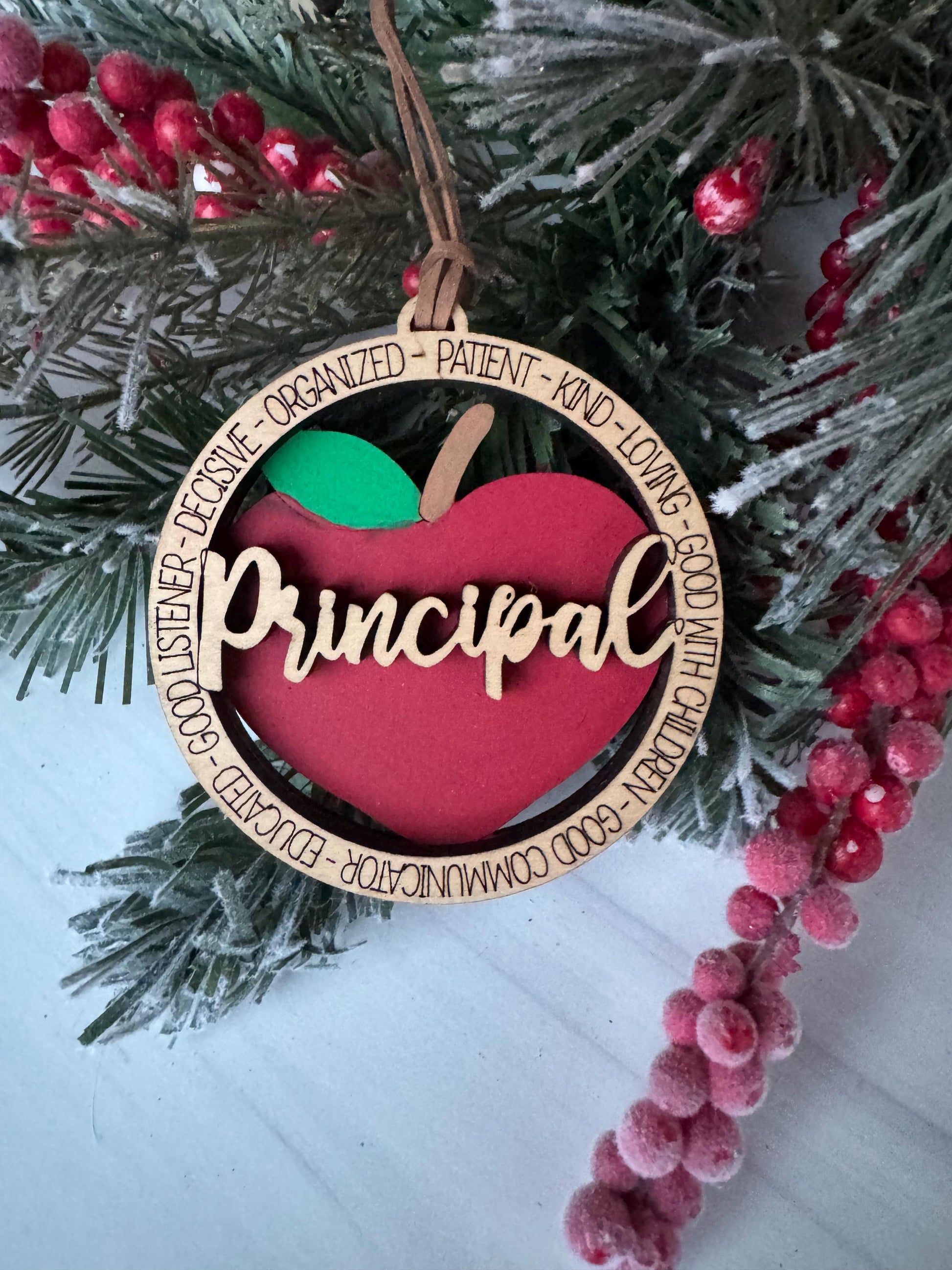 A red ornament with the word &quot;Principal&quot; on it, hanging on a pine branch with red berries.