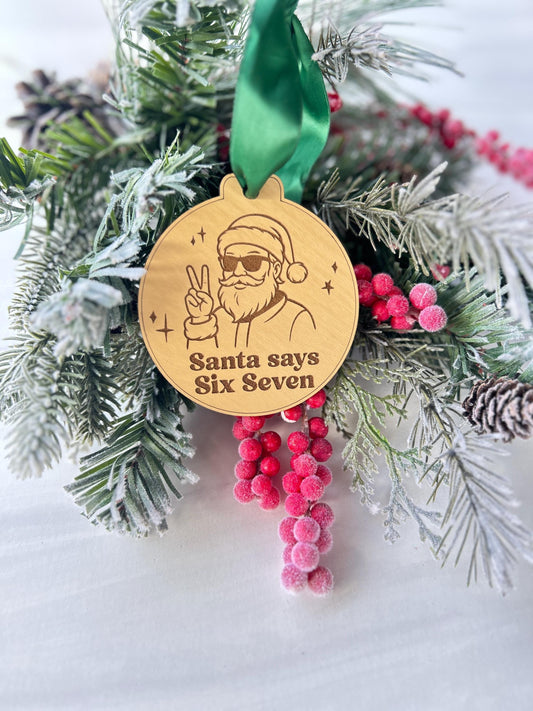A wooden ornament with a Santa Claus figure and the text &quot;Santa says Six Seven&quot; is hanging on a pine branch with pine cones and red berries.