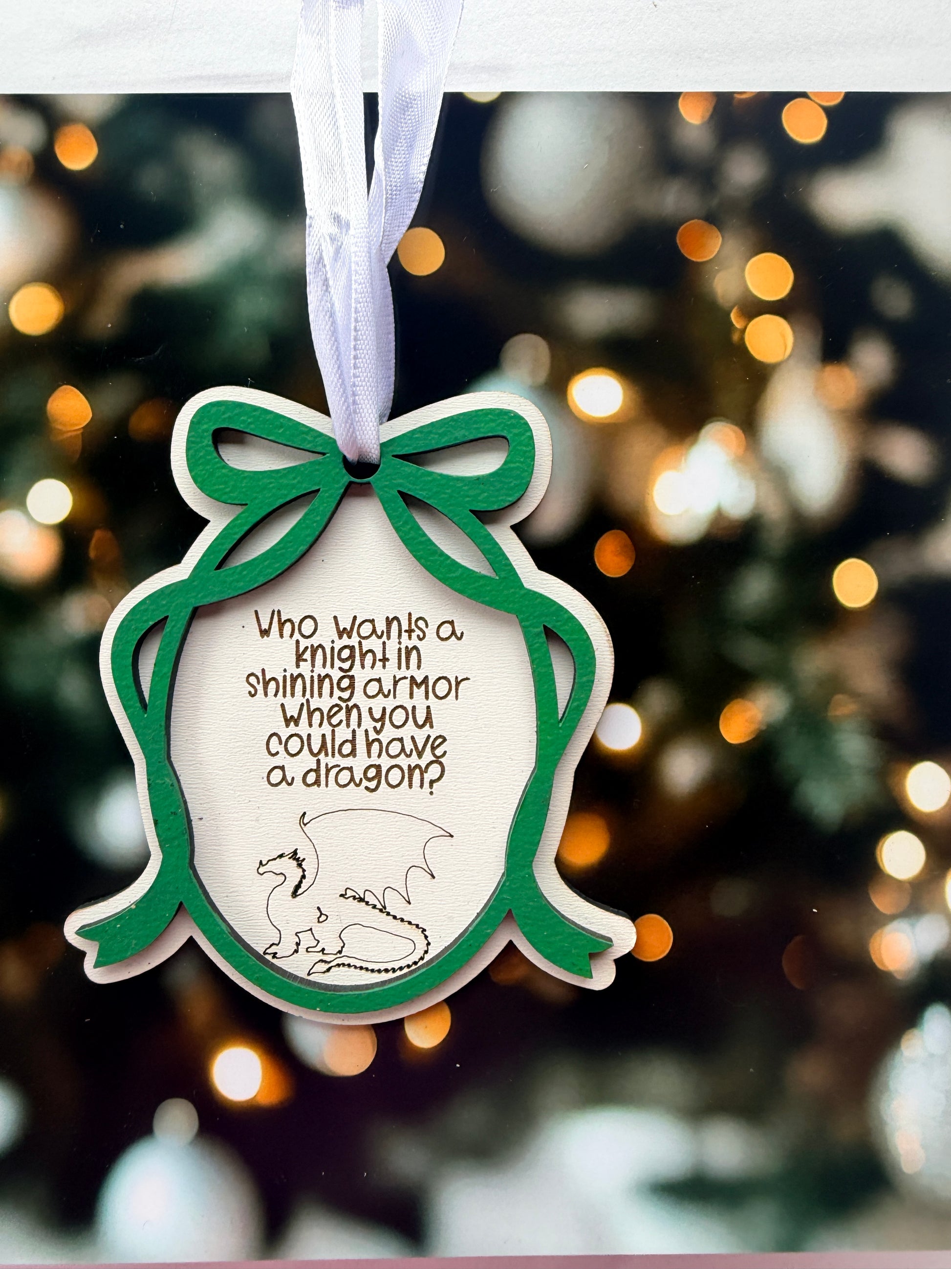 A wooden ornament with a dragon design and a green ribbon is hanging against a blurred background with Christmas lights.