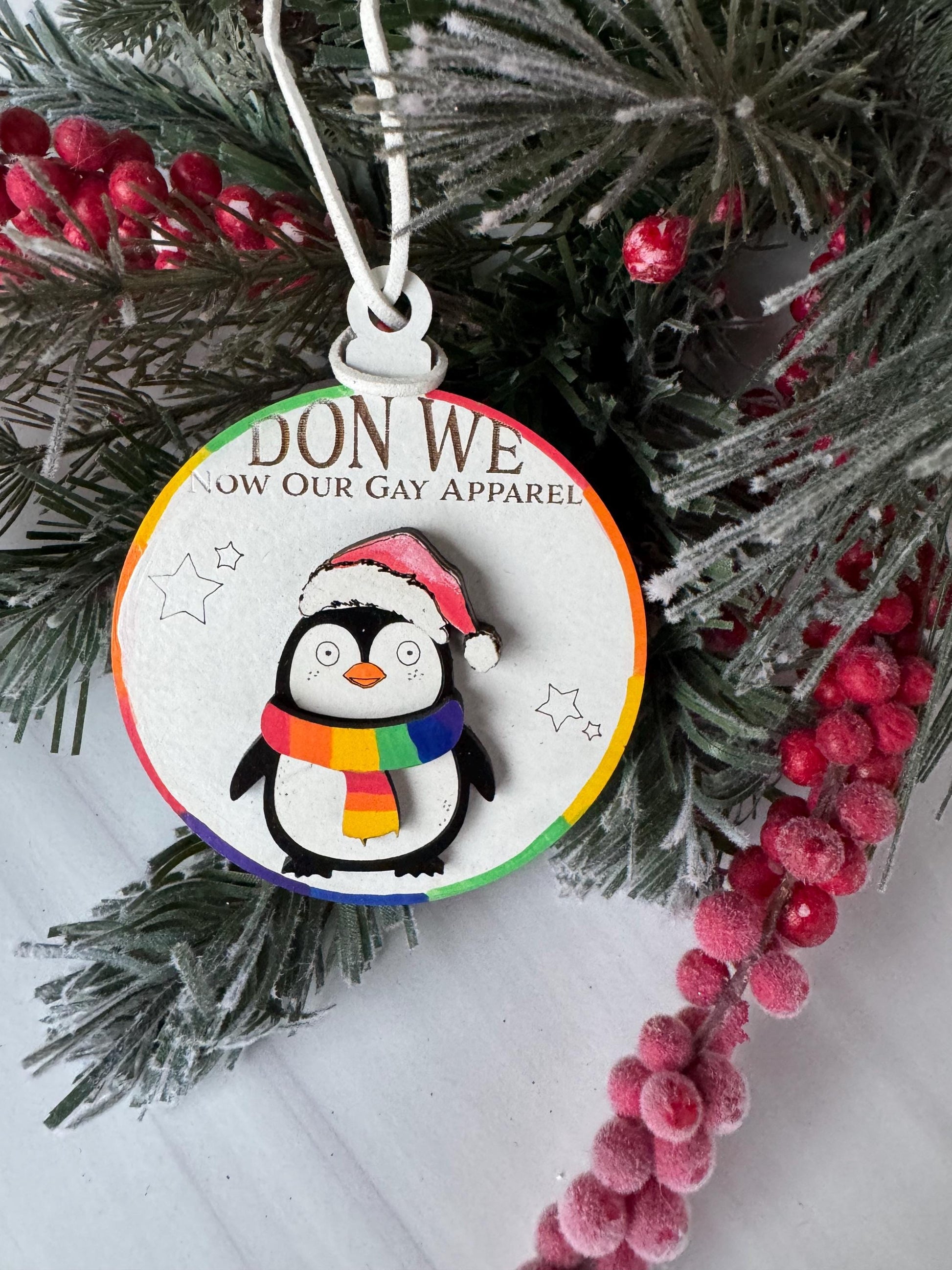 A Christmas ornament with a penguin wearing a rainbow-colored scarf, surrounded by pine branches and red berries.
