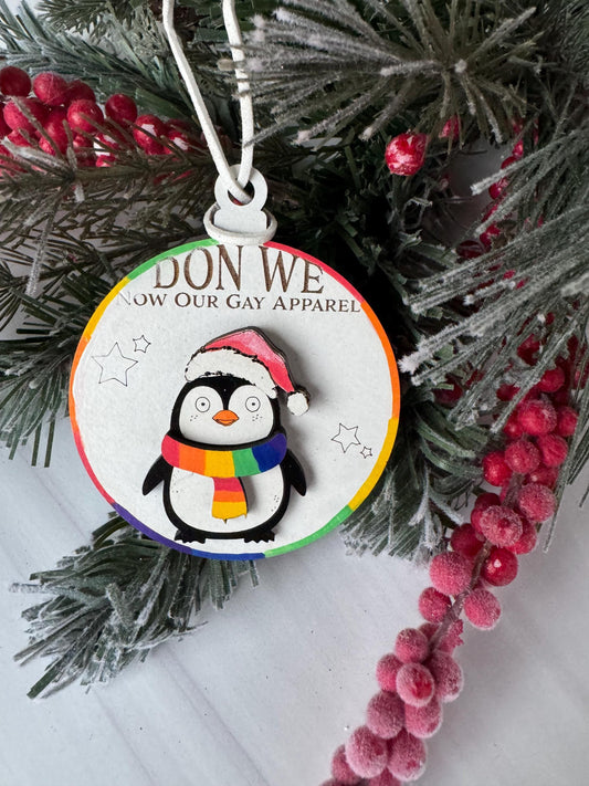 A Christmas ornament with a penguin wearing a rainbow-colored scarf, surrounded by pine branches and red berries.