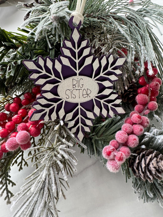 A purple snowflake ornament with the word "Big Sister" written on it, surrounded by pine branches, berries, and pine cones.