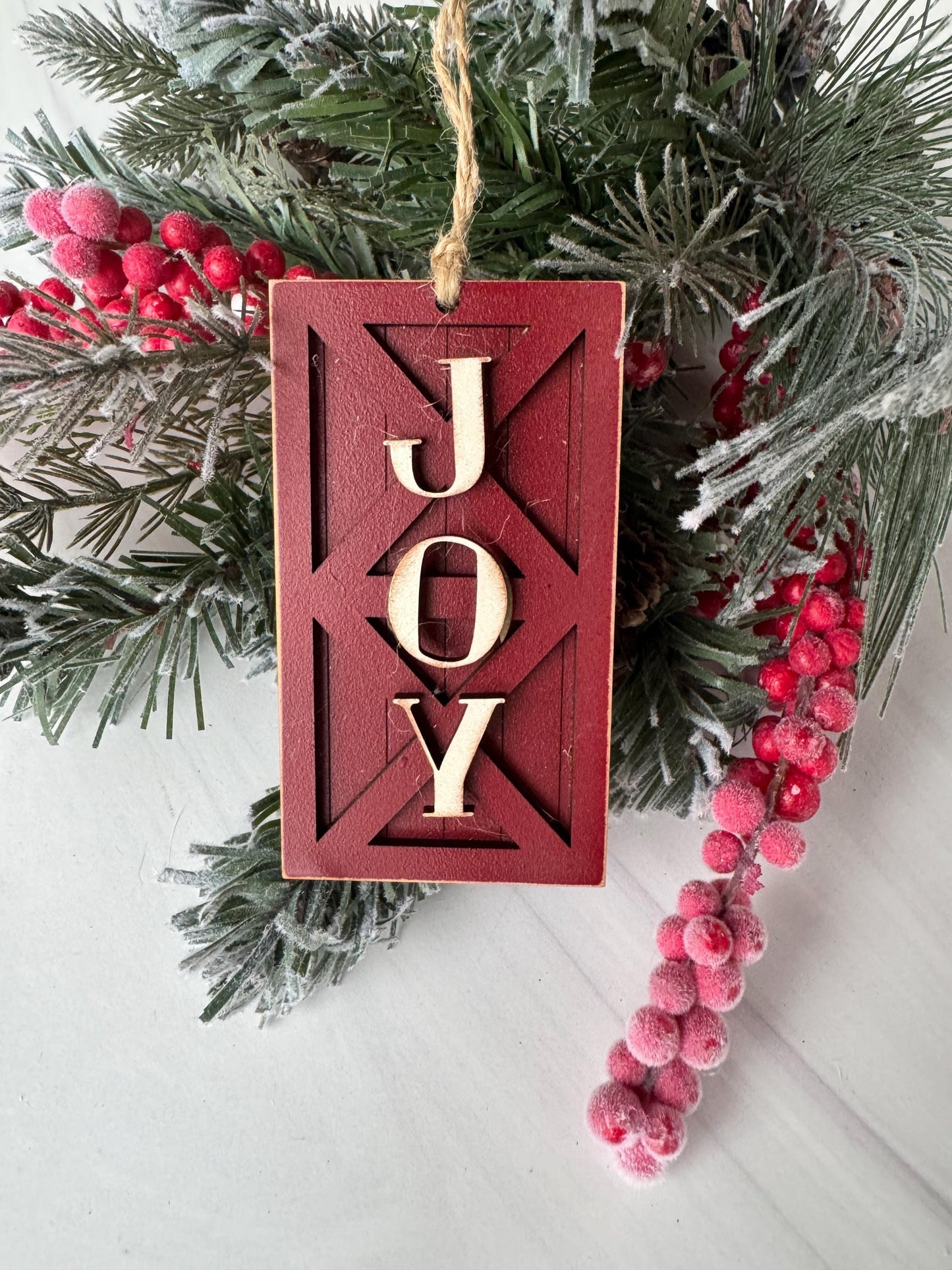 A red wooden ornament with the word &quot;Joy&quot; carved into it, surrounded by pine branches and red berries, creating a festive holiday scene.