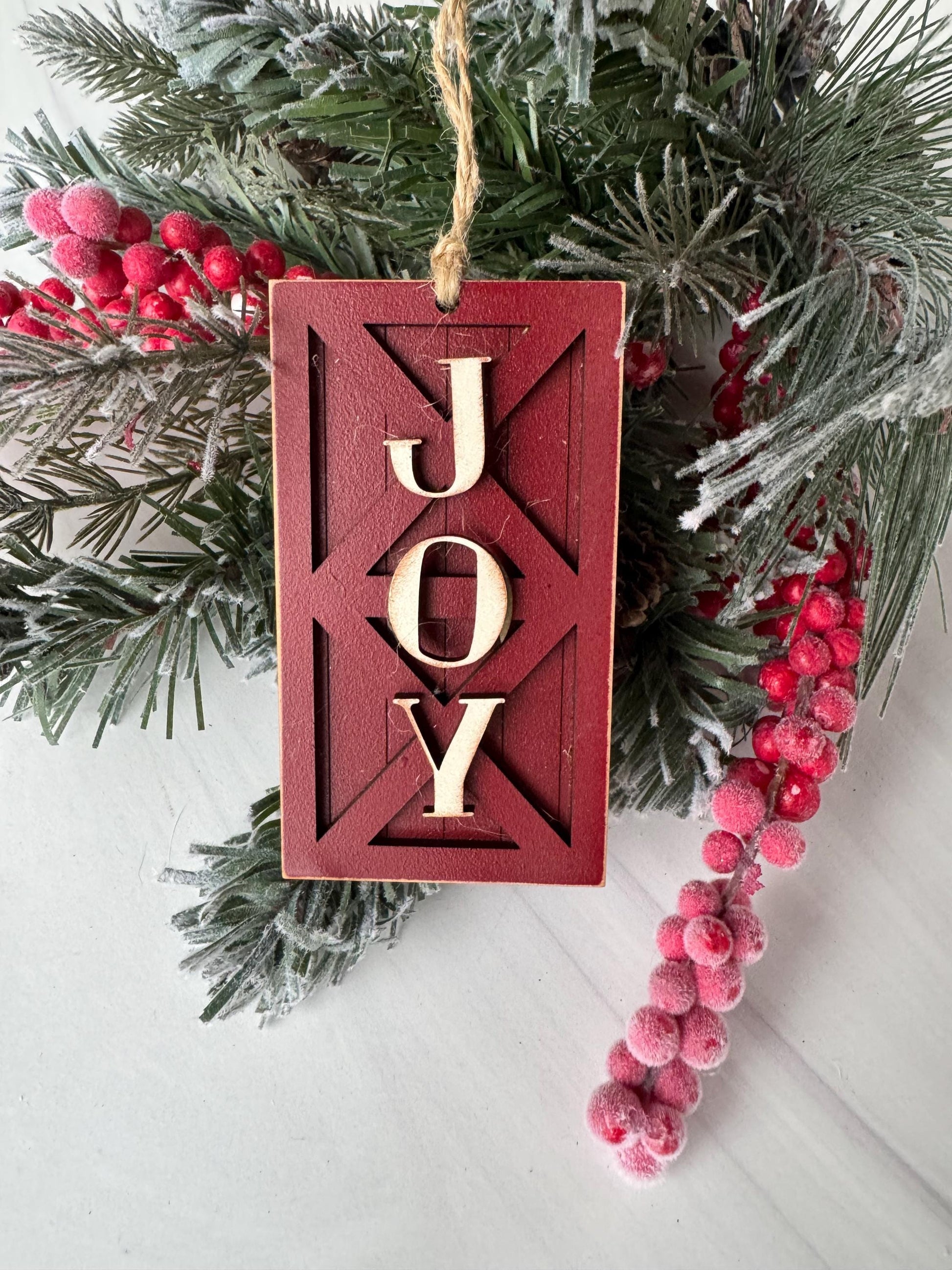 A red wooden ornament with the word &quot;Joy&quot; carved into it, surrounded by pine branches and red berries, creating a festive holiday scene.