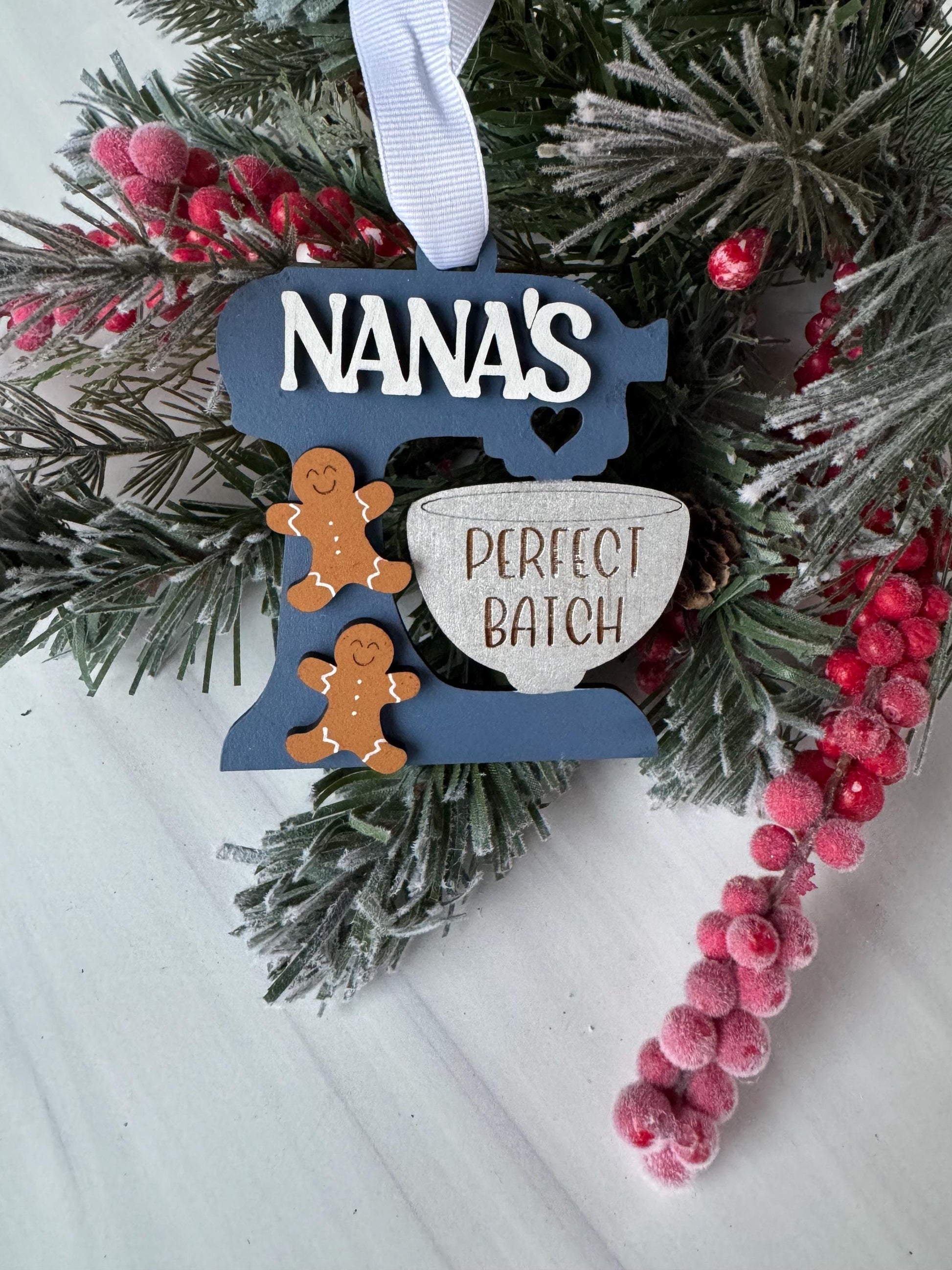 A Christmas ornament with the word "NANA's" written on it, surrounded by pine branches, red berries, and a white ribbon.