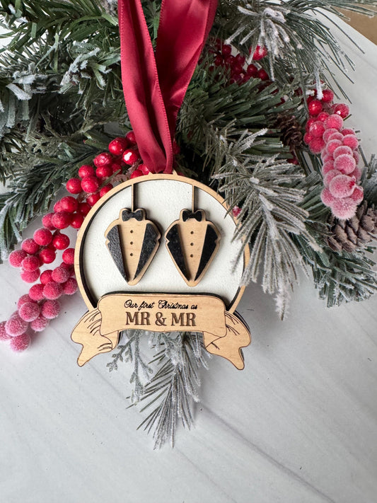 A decorative ornament with a ribbon and a pair of black tuxedo-shaped ornaments, surrounded by pine branches, red berries, and a red ribbon.