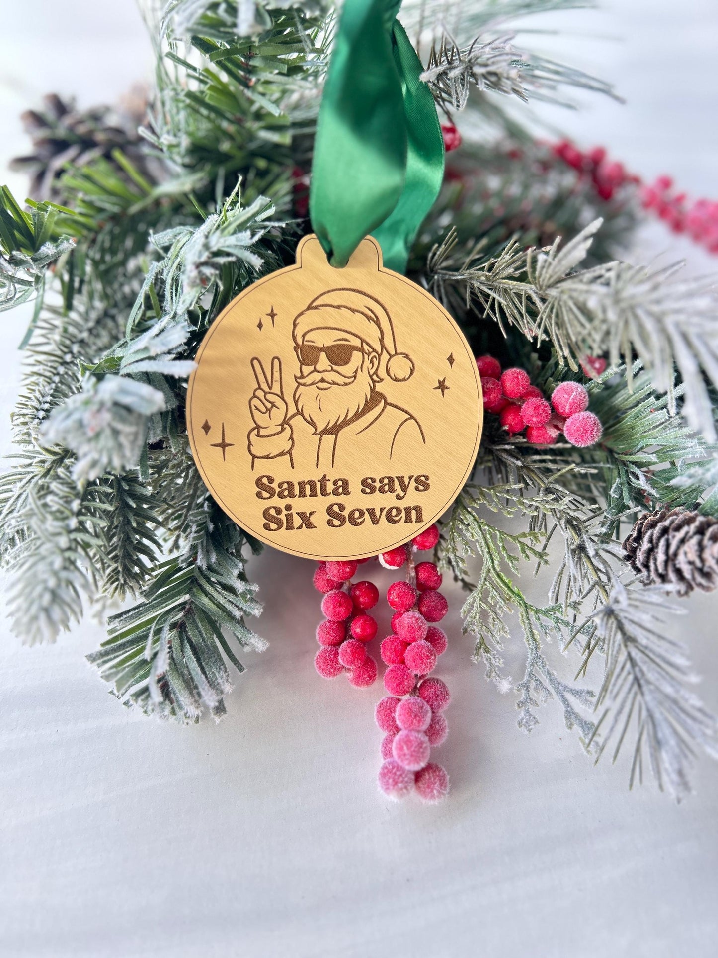 A wooden ornament with a Santa Claus figure and the text "Santa says Six Seven" is hanging on a pine branch with pine cones and red berries.