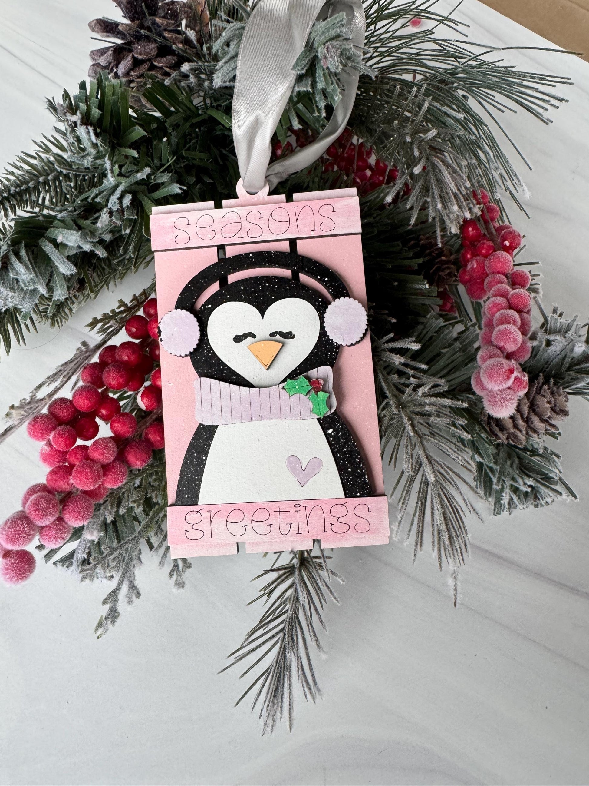 A pink and white penguin ornament is displayed on a white surface, surrounded by pine branches, red berries, and a white ribbon.