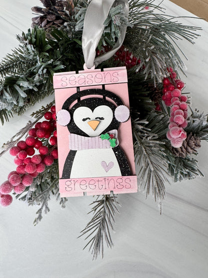 A pink and white penguin ornament is displayed on a white surface, surrounded by pine branches, red berries, and a white ribbon.
