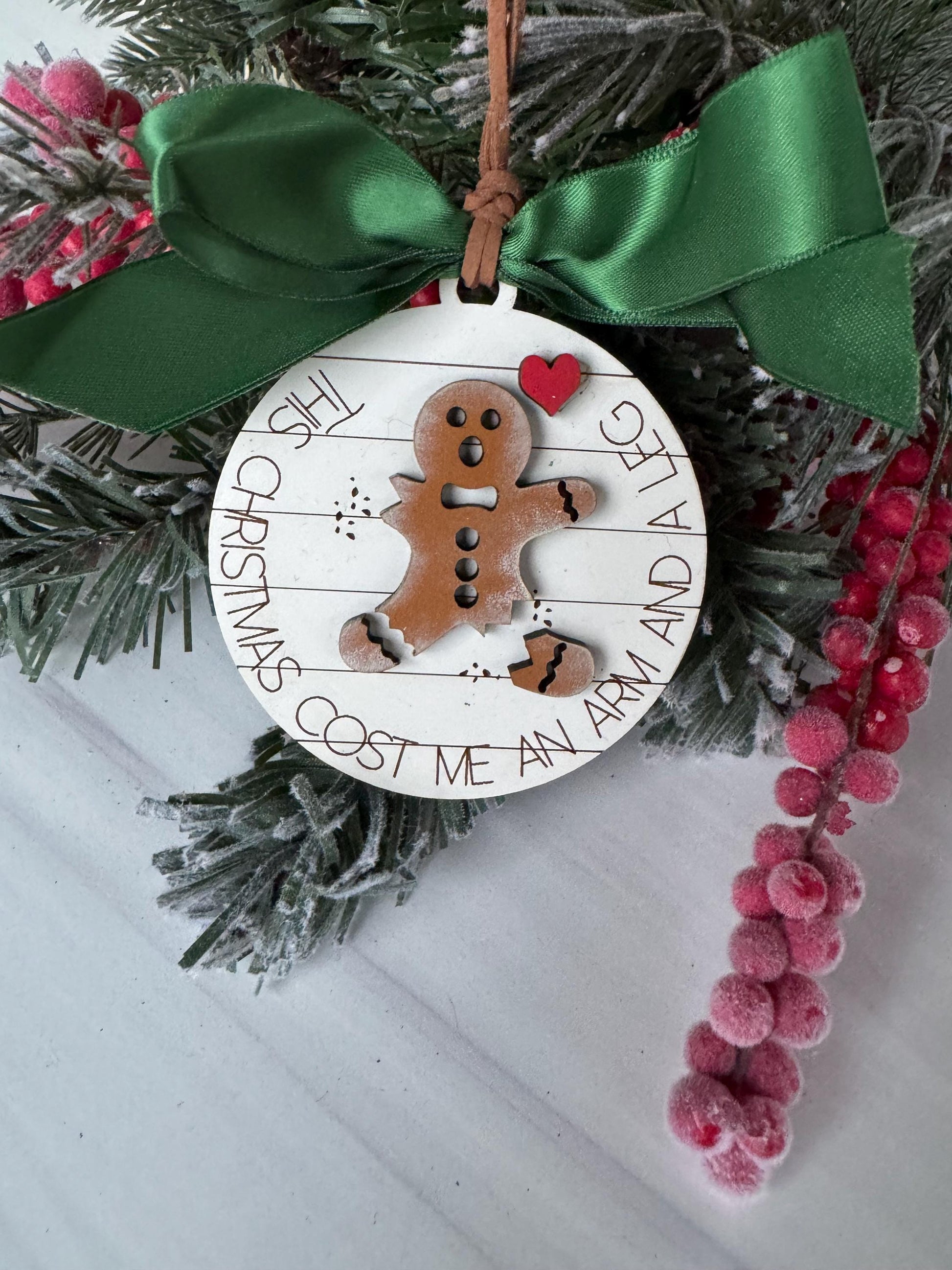 A Christmas ornament with a gingerbread man figure and a green ribbon, placed on a snowy surface with red berries in the background.