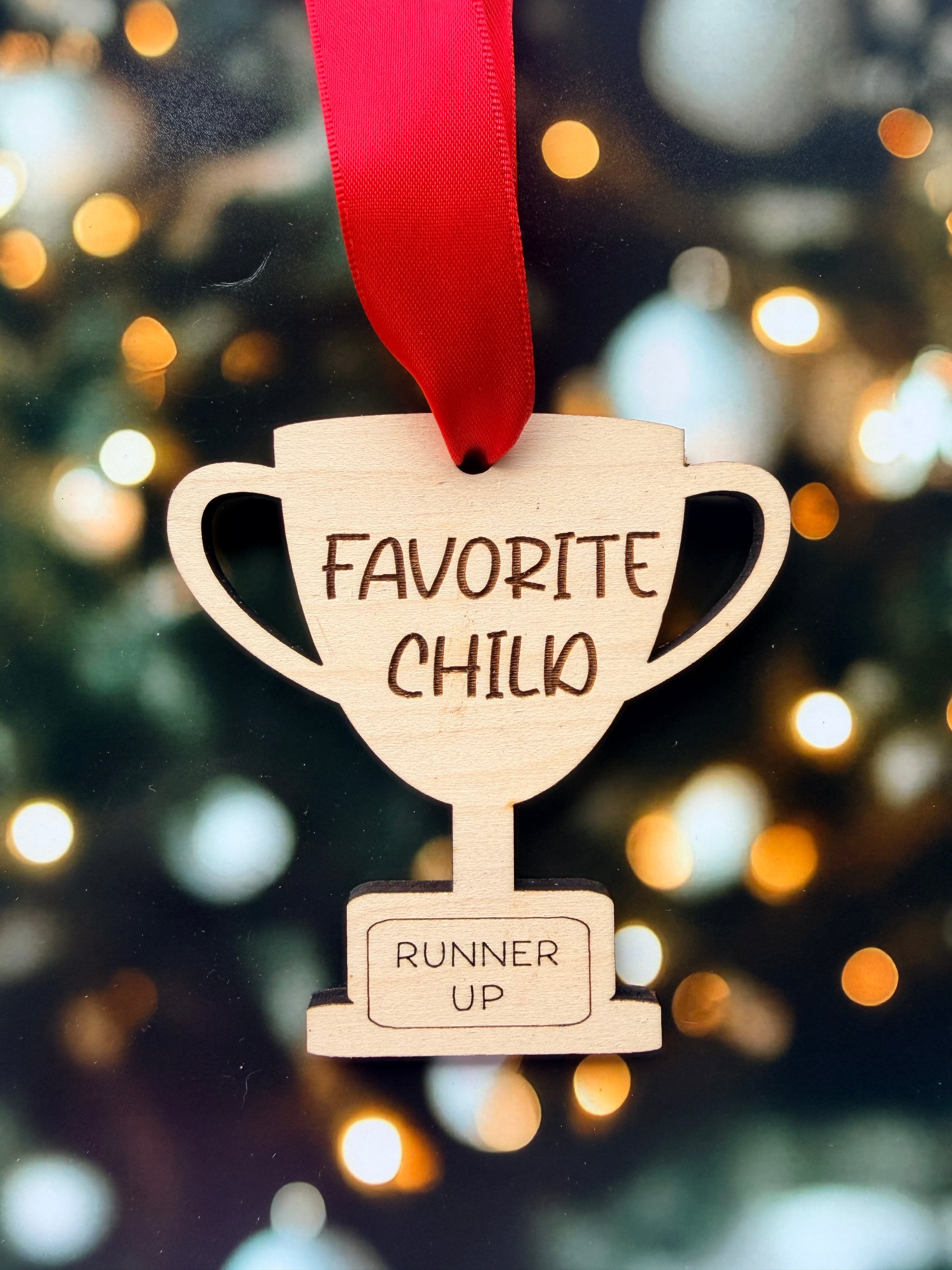 A wooden trophy-shaped ornament with a red ribbon attached, featuring the text "FAVORITE CHILD" and "RUNNER UP" on it.