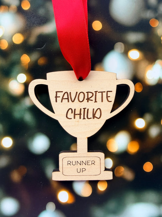 A wooden trophy-shaped ornament with a red ribbon attached, featuring the text "FAVORITE CHILD" and "RUNNER UP" on it.