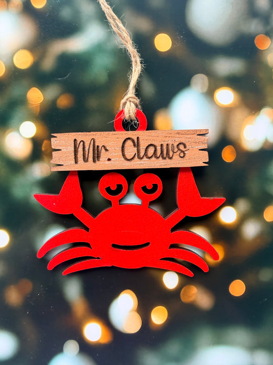 A red wooden ornament shaped like a crab with the name "Mr. Claws" written on it, hanging on a string against a background of blurred lights.