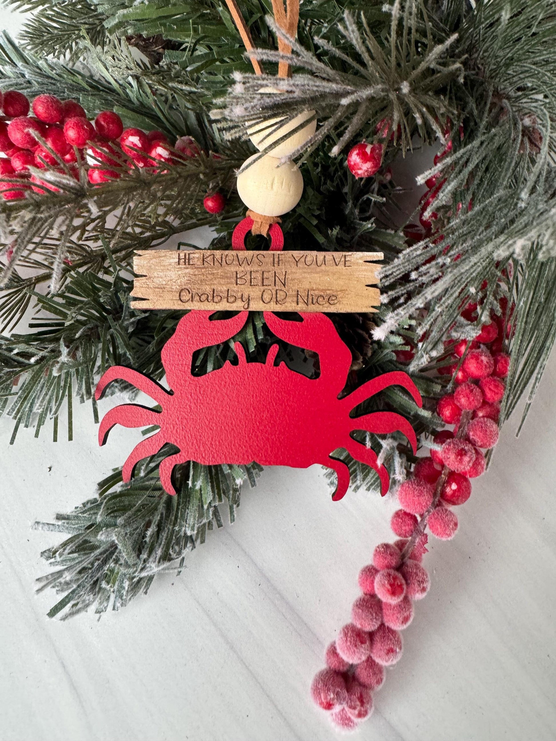 A red crab ornament is displayed among a wreath of pine branches and red berries, with a wooden sign featuring a message attached to it.